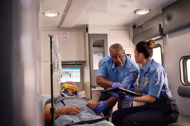 Two EMTs treating a patient on a stretcher in the back of an ambulance, one holding a tablet