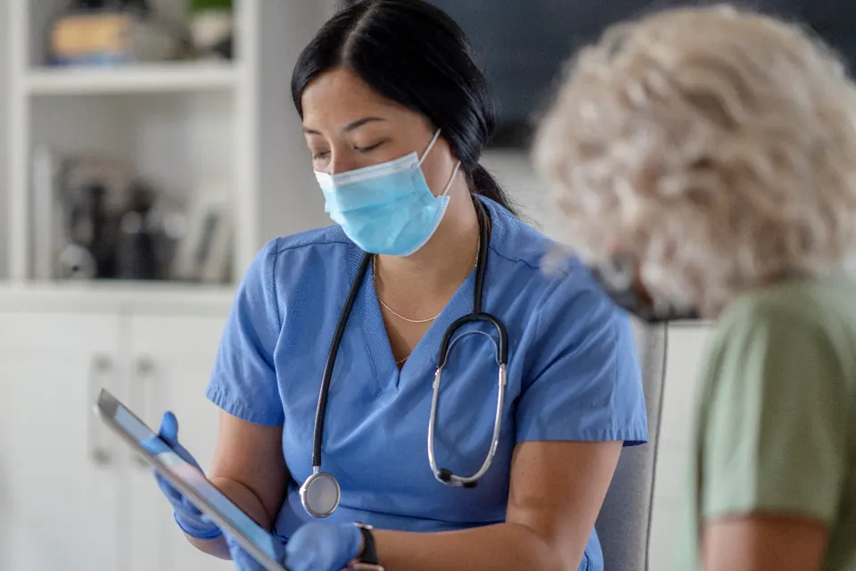 A masked clinician in scrubs holding a tablet beside a seated older patient